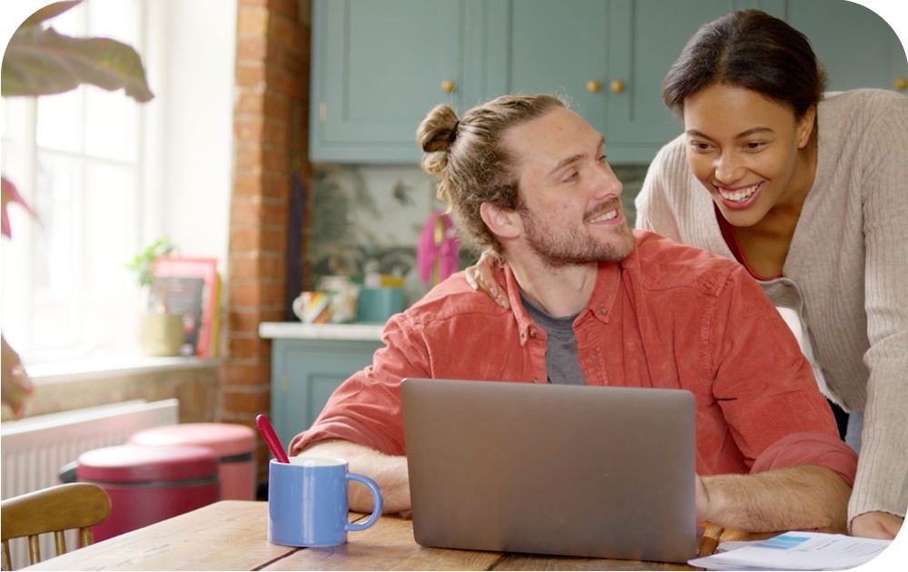 A couple gathered around rustic wooden table smiling and discussing contents on a laptop next to a coffee cup. 