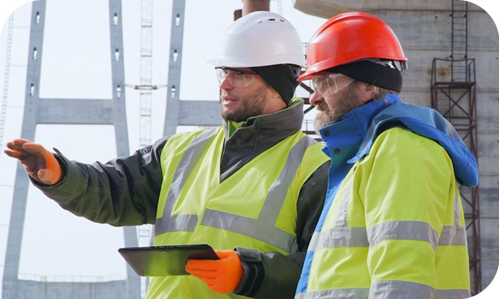 Two field workers in hard hats and neon yellow vests look out across the site and discuss updates with a tablet between them