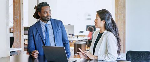 Two coworkers talk and work together in a work setting in front of a laptop