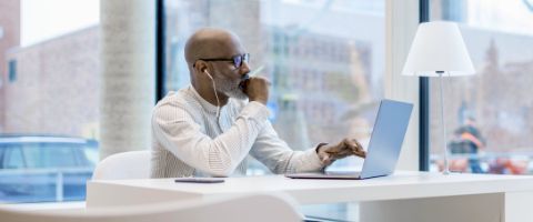 Man sitting with laptop open at office desk next to city scaped window in brightly lit conference room.  