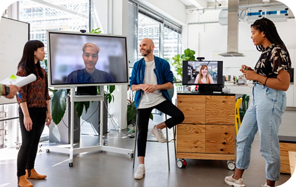 Three people in casual attire standing in an office setting with others on a video call