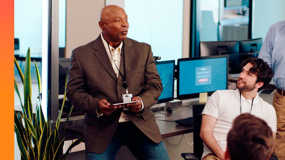 Business professional stands and speaks to people in open office while coworkers listen. 