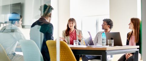 Five people sitting at a conference table while talking and working with laptop devices