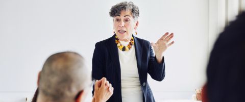 Woman in a dark colored jacket standing while addressing people sitting at a table in front of her